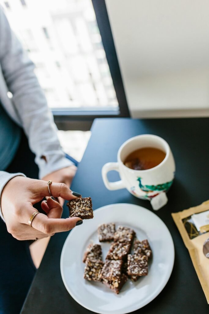 Hand holding chocolate snack near a hot beverage, with more brownies on a plate.